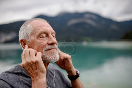 Photo for Senior listening music while running by lake in nature. Elderly man exercising to stay healthy, vital, enjoying physical activity, relaxation outdoors. - Royalty Free Image