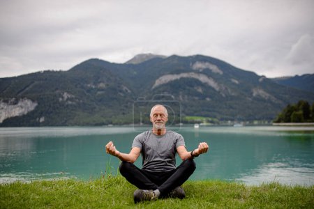 Téléchargez les photos : Homme âgé faisant du yoga en plein air, méditant au bord du lac dans les montagnes. Un sportif âgé passe des vacances actives dans la nature. Pensionné étirement après l'entraînement en plein air - en image libre de droit