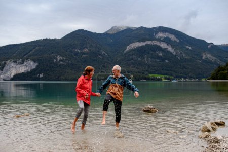Active elderly couple hiking together in spring mountains, walking in cold fresh water in mountain lake. Senior tourists enjoying nature.
