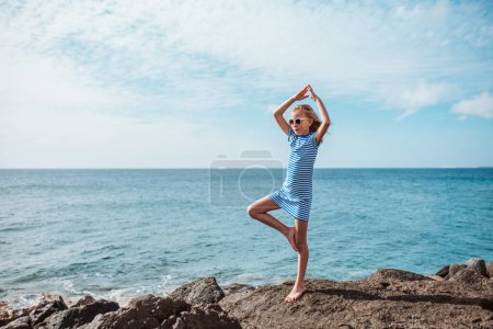 Young girl standing on one leg on rock. Blonde girl in striped dress enjoying vacation in Canary Islands. Concept of beach summer vacation with kids.
