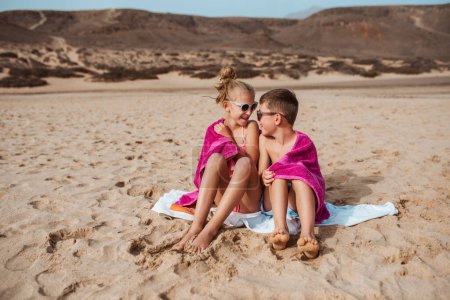 Téléchargez les photos : Frères et sœurs assis sur la plage, enveloppés dans une serviette après avoir nagé dans la mer. Concept de vacances d'été à la plage familiale avec enfants. - en image libre de droit