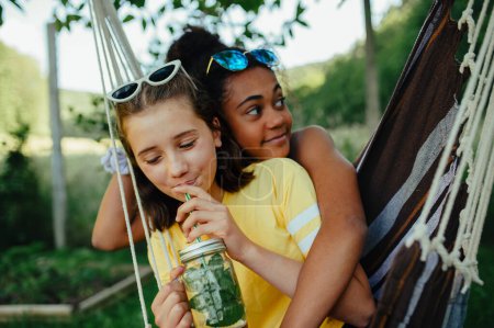 Téléchargez les photos : Yong adolescentes amis à l'extérieur dans le jardin, balançant sur la balançoire, boire de la limonade à partir de verre de bocal, pendant la chaude soirée d'été. - en image libre de droit