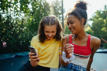 Téléchargez les photos : Les adolescentes amies passent du temps dehors dans le jardin, riant. Assis sur le trampoline, défilant sur smartphone, les médias sociaux, pendant la chaude journée d'été. - en image libre de droit