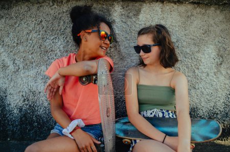 Téléchargez les photos : Jeune adolescente meilleures amies avec des planches à roulettes passer du temps en ville pendant la chaude journée de vacances d'été. Assis près d'un mur de béton. - en image libre de droit