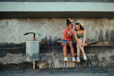 Téléchargez les photos : Jeune adolescente meilleures amies avec des planches à roulettes passer du temps à l'extérieur en ville pendant la chaude journée de vacances d'été. Assis sur un mur de béton, parlant, partageant un secret. - en image libre de droit
