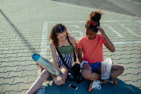 Young teenager girl best friends with skateboards spending time outdoors in city during warm summer holiday day. Sitting on parking lot, talking.