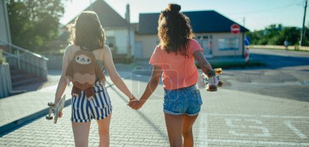 Photo for Rear view of young teenager girl best friends with skateboards spending time outdoors in city during warm summer holiday day. Banner with copy space. - Royalty Free Image