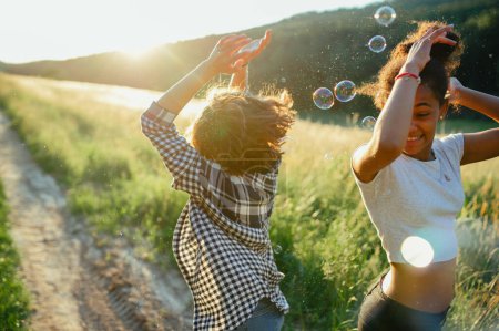 Photo for Cheerful young teenager girl best friends spending time in nature, during sunset. Girls blowing bubbles and popping them. - Royalty Free Image