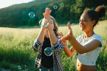 Foto de Jovencita alegre adolescente mejores amigos pasar tiempo en la naturaleza, durante la puesta del sol. Chicas soplando burbujas y haciendo estallar ellos. - Imagen libre de derechos