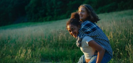 Photo for Cheerful young teenager girl best friends spending time in nature, during sunset. Girls on walk, carrying each other on back. - Royalty Free Image