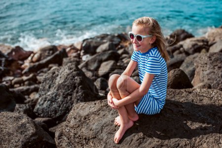 Beautiful girl sitting on big rocks by sea. Blonde girl in dress enjoying vacation in Canary Islands. Concept of beach summer vacation with kids.