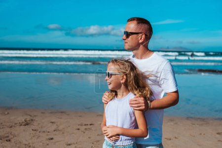 Foto de Padre e hija, de pie en la playa. Papá y niña disfrutando de la playa de arena, mirando el mar cristalino en las Islas Canarias. Concepto de vacaciones de verano en la playa con niños. - Imagen libre de derechos