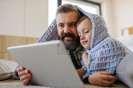 Téléchargez les photos : Petit garçon regardant un film de dessin animé sur la tablette avec son père, couché sous la couverture sur le sol dans la chambre des enfants. Papa expliquant la technologie au fils, littératie numérique pour les enfants. - en image libre de droit