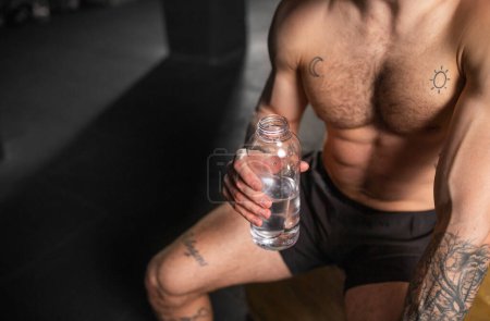 Photo for Close up of man resting after exercise, drinking water from bottle, sitting, wearing short with muscular bare chest. Routine workout for physical and mental health. - Royalty Free Image