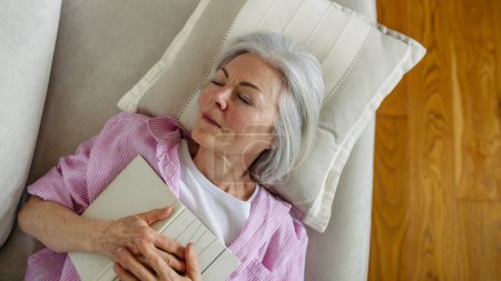 Foto de Vista superior de una hermosa mujer madura dormida con libro. Actividad de fin de semana para mujeres mayores, relajarse en casa. - Imagen libre de derechos