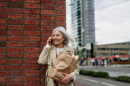 Foto de Mujer madura haciendo una llamada, volviendo a casa de Supermaket con gorceries. Hermosa mujer mayor con pelo gris de pie en la calle de la ciudad. - Imagen libre de derechos