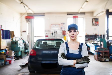 Photo for Female auto mechanic repairing, maintaining a car. Beautiful woman standing in a garage, wearing blue coveralls. Female automotive service technican. - Royalty Free Image