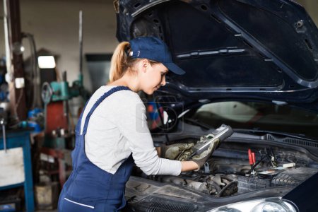 Photo for Female auto mechanic using diagnostic tool, scanner, running diagnostic on car. Beautiful woman working in a garage, wearing blue coveralls. Performing diagnostic test. - Royalty Free Image