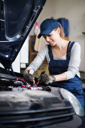 Photo for Female auto mechanic repairing, maintaining a car battery. Beautiful woman working in a garage, wearing blue coveralls. Female automotive service technican. - Royalty Free Image