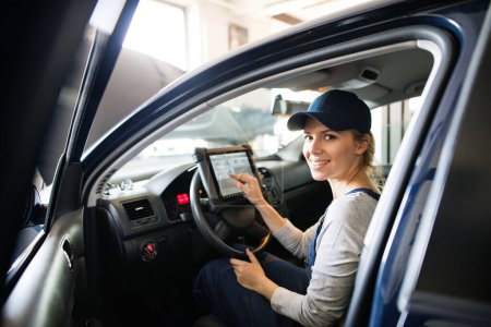 Photo for Female auto mechanic using diagnostic tool, scanner, running diagnostic on car. Beautiful woman sitting inside of car, performing diagnostic test. Woman working in a garage, wearing blue coveralls. - Royalty Free Image