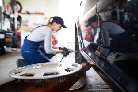 Automechanikerin wechselt die Reifen im Autoservice. Schöne Frau kniet auf einem Rad in einer Garage und trägt blaue Overalls. Kfz-Servicetechnikerin.