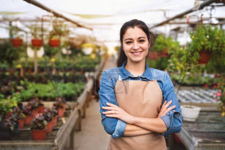 Photo for Small greenhouse business. Businesswoman selling flowers and seedlings, standing in the middle of plants, looking at camera, smiling. Offering wide range of plants during spring gardening season. - Royalty Free Image