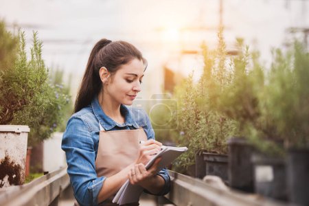 Small greenhouse business. Female gardener inspecting flowers and seedlings, standing in the middle of plants with clipboard, smelling them. Offering wide range of plants during spring gardening