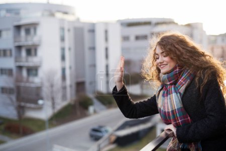 Foto de Hermosa mujer con el pelo rizado de pie en una terraza, saludando a alguien en la calle. Sol de la mañana durante el frío día de primavera. - Imagen libre de derechos