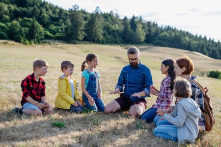 Children and teachers sitting on grass on meadow playing clapping game. Dedicated teachers during outdoor active education teaching about ecosystem, ecology and nature.