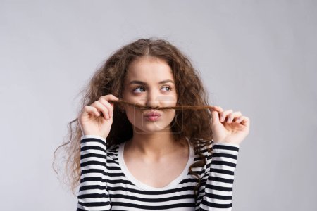 Photo for Portrait of a gorgeous teenage girl with curly hair, holding lock of hair as a moustache. Studio shot, white background with copy space. - Royalty Free Image