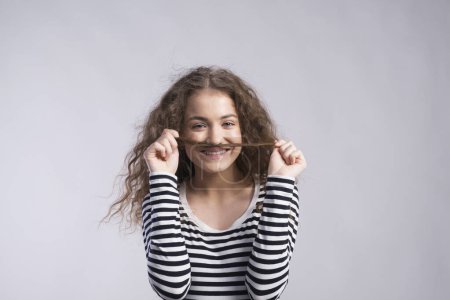 Téléchargez les photos : Portrait d'une magnifique adolescente aux cheveux bouclés, tenant une mèche de cheveux comme une moustache. Plan studio, fond blanc avec espace de copie. - en image libre de droit