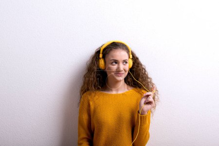 Photo for Portrait of a gorgeous teenage girl with curly hair, listening music via yellow headphones. Studio shot, white background with copy space. - Royalty Free Image
