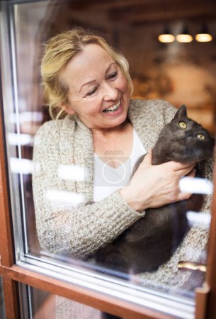 Hermosa mujer madura en casa, sosteniendo, acariciando a su gato, mirando a través de la ventana. Mujer mayor viviendo sola, disfrutando de un fin de semana tranquilo.