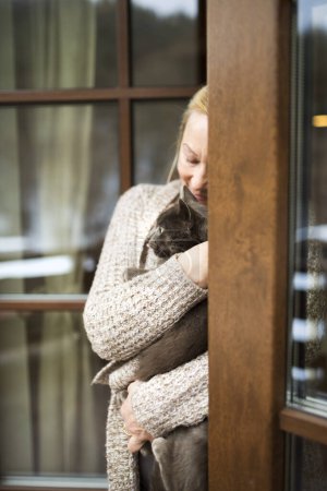 Foto de Hermosa mujer madura en casa, sosteniendo, acariciando a su gato, mirando a través de la ventana. Mujer mayor viviendo sola, disfrutando de un fin de semana tranquilo. - Imagen libre de derechos