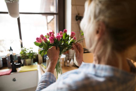 Foto de Hermosa mujer madura organizando un ramo de rosas en un jarrón, un hobby y relajación. Mujer mayor viviendo sola, disfrutando de un fin de semana tranquilo. - Imagen libre de derechos
