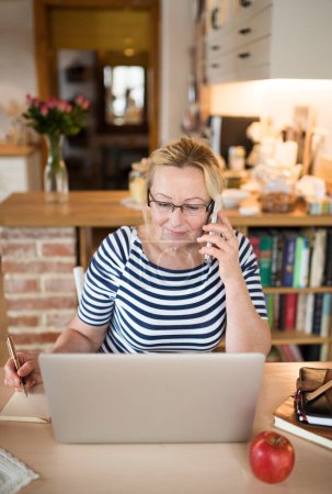 Older woman working from homeoffice, typing on laptop in the kitchen and making phone calls. The retiree earning extra money during retirement.
