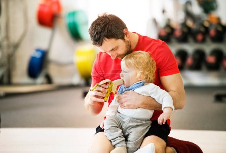 Photo for Portrait of new dad on group exercise class in gym, feeding baby with banana. Parents staying active while boding with babies. Baby movement class. - Royalty Free Image