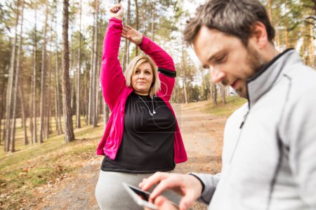 Overweight woman stretching befor run, personal trainer checking her profile, performance on tablet. Exercising outdoors for people with obesity, support from male fitness coach.