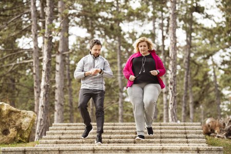Overweight woman running down the stairs, personal trainer checking her performance. Exercising outdoors for people with obesity, support from friend or fitness coach.