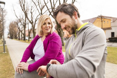 Overweight woman resting after run, personal trainer checking her running speed on smartwatch. Exercising outdoors for people with obesity, support from male fitness coach.