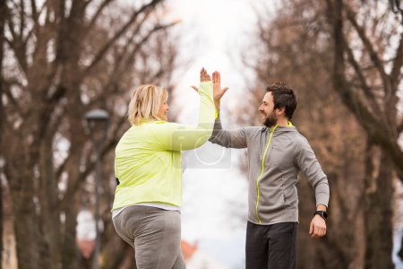 An overweight woman running in nature with friend, high five. Exercising outdoors for people with obesity, support from friend or fitness coach.