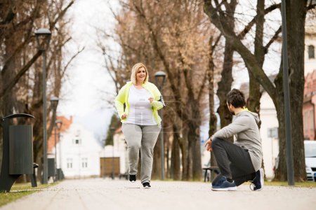 Photo for Overweight woman running in city park, personal trainer checking her performance. Exercising outdoors for people with obesity, support from friend or fitness coach. - Royalty Free Image