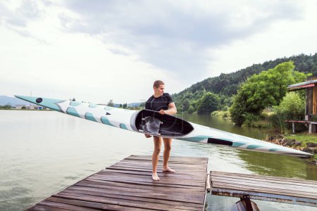 Foto de El joven piragüista lleva canoa y remo, va al agua, camina sobre un muelle de madera. Concepto de piragüismo como deporte dinámico y aventurero. - Imagen libre de derechos