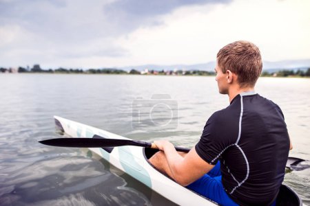 Canoeist man sitting in canoe holding paddle, in water. Concept of canoeing as dynamic and adventurous sport. Rear view, sportman looking at water surface, paddling.