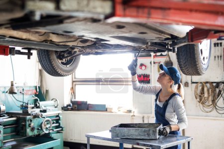 Photo for Female auto mechanic elevating car on car, automotive lift, changing oil. Beautiful woman working in a garage, wearing blue coveralls. Female automotive service technican. - Royalty Free Image