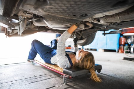 Photo for Female auto mechanic lying on mechanic creeper under car, inspecting and repairing vehicle. Beautiful woman working in a garage, wearing blue coveralls. Female automotive service technican. - Royalty Free Image