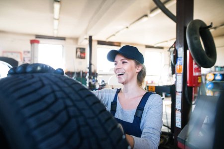 Automechanikerin wechselt die Reifen im Autoservice. Schöne Frau mit Reifen in einer Garage, in blauen Overalls. Reifenmontage. Kfz-Servicetechnikerin.