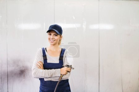 Foto de Mujer pintor de coches, vehículos de paingting en taller de carrocería. Mujer joven sosteniendo pistola, mirando a la cámara y sonriendo. - Imagen libre de derechos