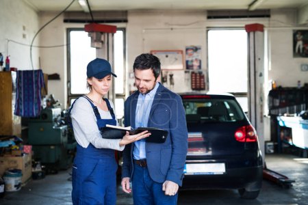 Automechanikerin im Gespräch mit dem Kunden, erklärt Reparatur, Problem. Termin für die jährliche Wartung. Schöne Frau, die in einer Garage arbeitet und blaue Overalls trägt.