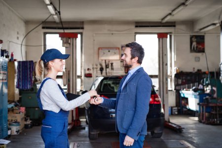 Photo for Female auto mechanic talking with customer, handing over keys of repaired car. Beautiful woman working in a garage, wearing blue coveralls. Female automotive service technican. - Royalty Free Image